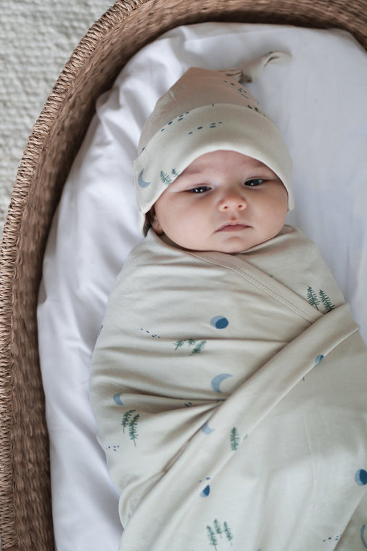 Baby laying in Moses Basket while wrapped in Midnight Forest Swaddle Blanket and wearing Midnight Forest Beanie, all designed by Burrow and Be