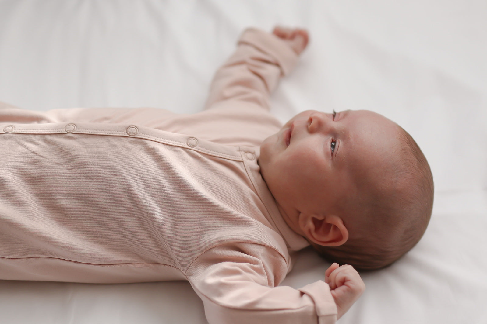 Baby lying on white sheets wearing Essential Sleep Suit in Dusty Rose, designed by Burrow and Be