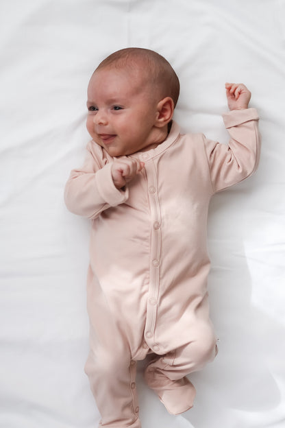 Baby smiling and laying on white sheets and wearing Essential Sleep Suit in Dusty Rose, designed by Burrow and Be