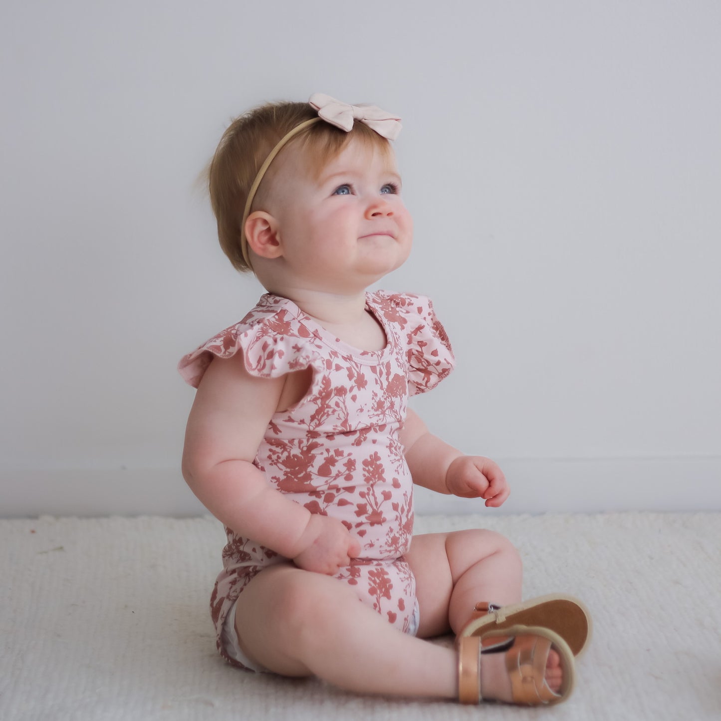 Baby girl wearing bow tie hair band, Flower Splash Flutter Bodysuit, Wander Sandals and Flower Splash Bike Shorts, all designed by Burrow and Be