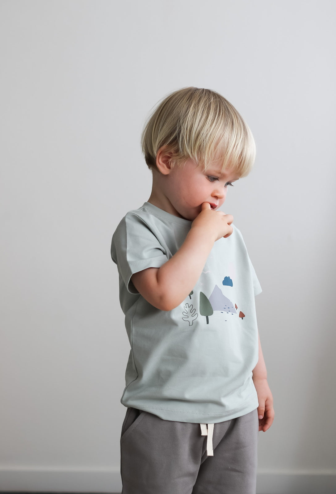 Baby boy standing against a white wall wearing Garden Treasures T-Shirt and Terry Knit Baby Shorts in Steel, all designed by Burrow and Be.