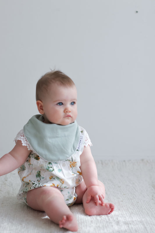 Baby wearing Burrow and Be Spring Melody Lace Sleeve Milly Romper with Pink Dribble Bib, in front of a white background