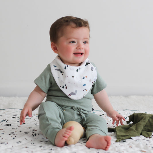 Baby sitting on Garden Treasure Quilted Mat, in front on a white wall while wearing Garden Treasure Dribble Bib, Pointelle Short Sleeve Bodysuit and Baby Pants in Riverstone, all designed by Burrow and Be