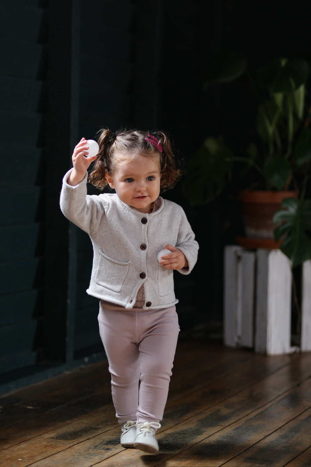 Baby girl holding two ping pong balls, wearing White Knit Cardigan and Essential Derby Rib Leggings - Lavender both designed by Burrow and Be