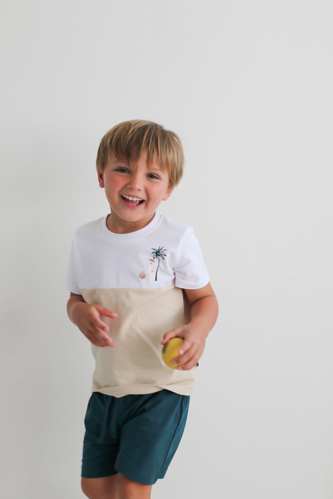 Boy holding a ball in hand and standing against a white wall while wearing Jungle Stamp Block T-Shirt, designed by Burrow and Be