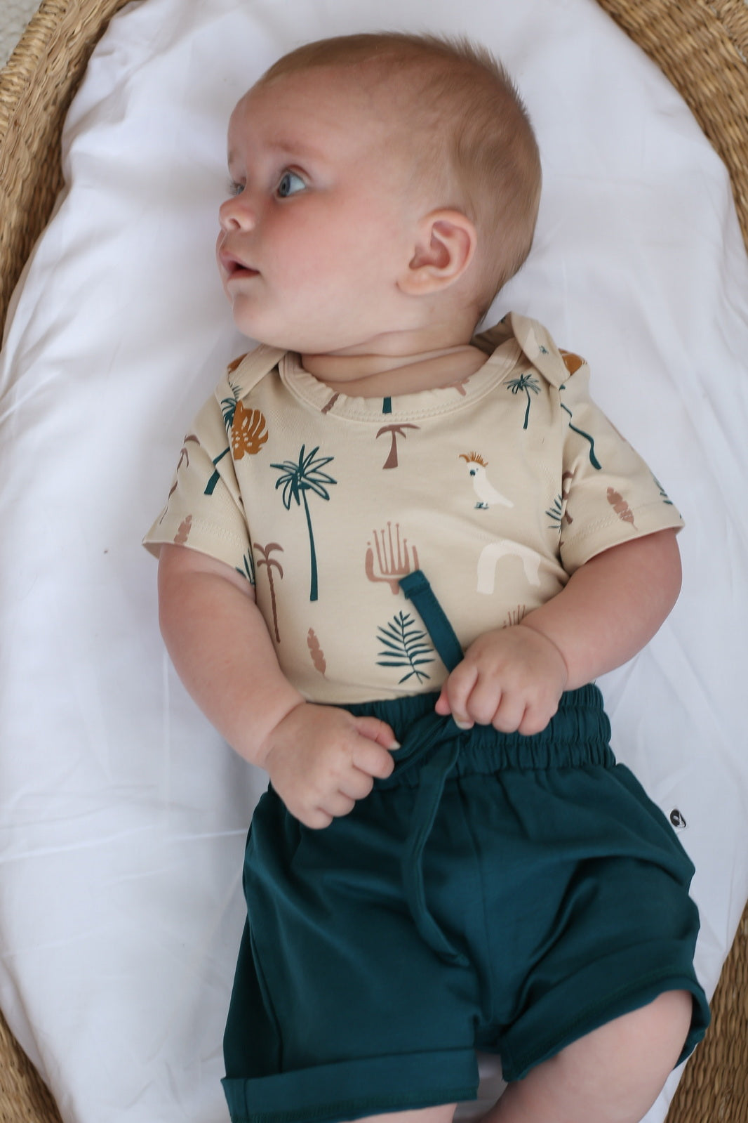 Baby laying in Moses Basket and wearing Leo Shorts in Deep Teal and Jungle Stamp Bodysuit, all designed by Burrow and Be