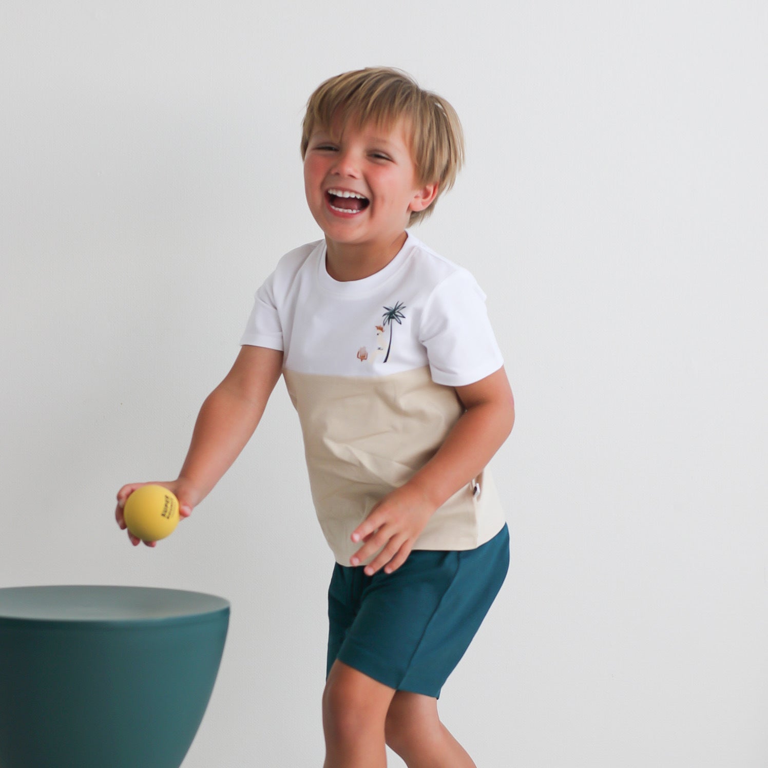 Boy holding a ball in hand and posing against a white background while wearing Jungle Stamp Block T-Shirt and Leo Shorts in Deep Teal, all designed by Burrow and Be