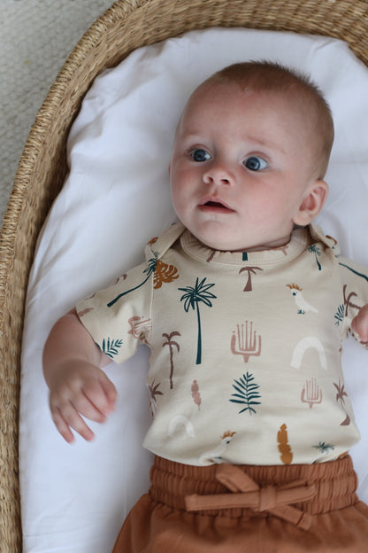 Baby staring at camera while laying in Moses Basket and wearing Leo Shorts in Tan and Jungle Stamp Bodysuit, all designed by Burrow and Be