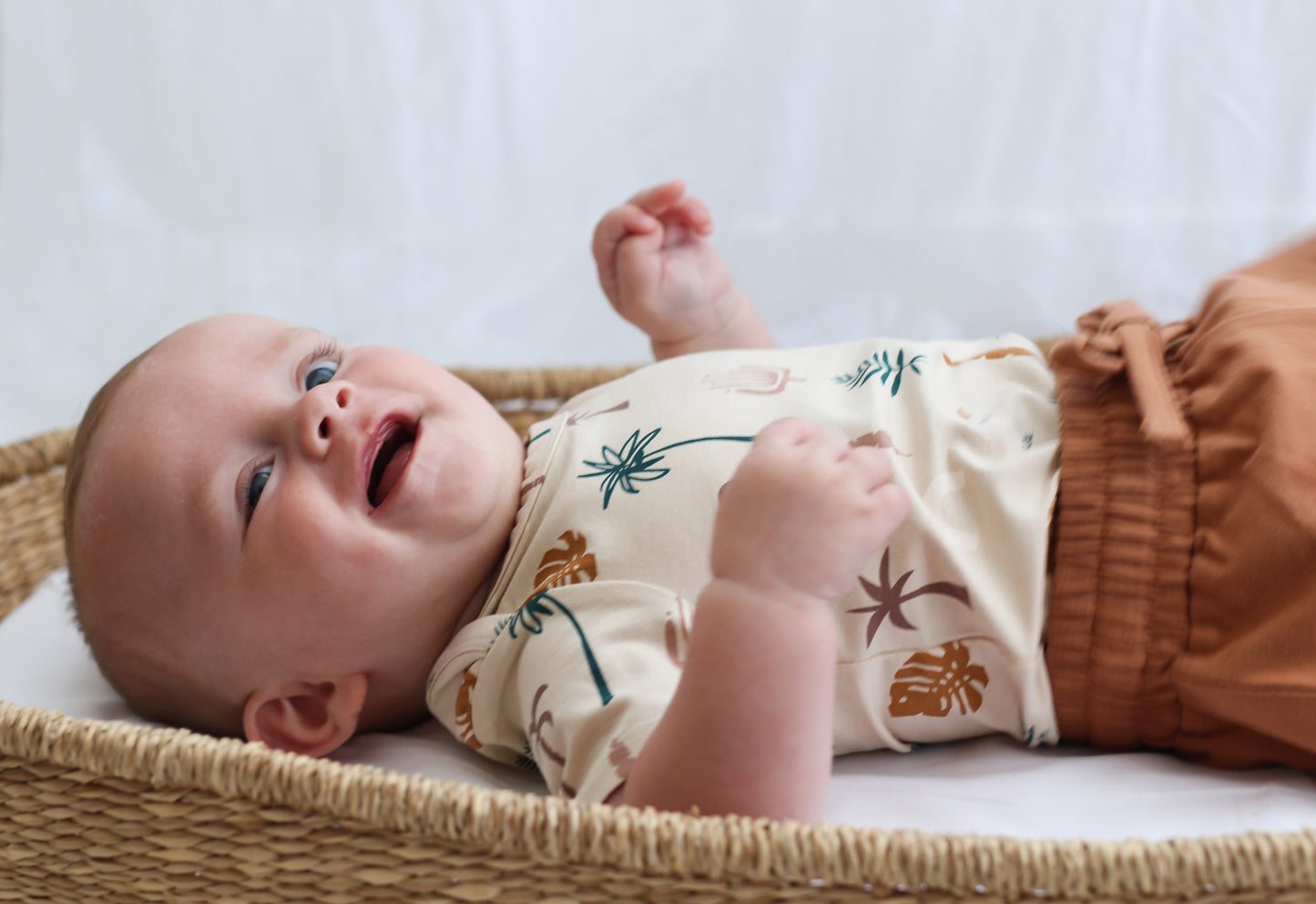 Baby staring at camera while laying in Moses Basket and wearing Leo Shorts in Tan and Jungle Stamp Bodysuit, all designed by Burrow and Be