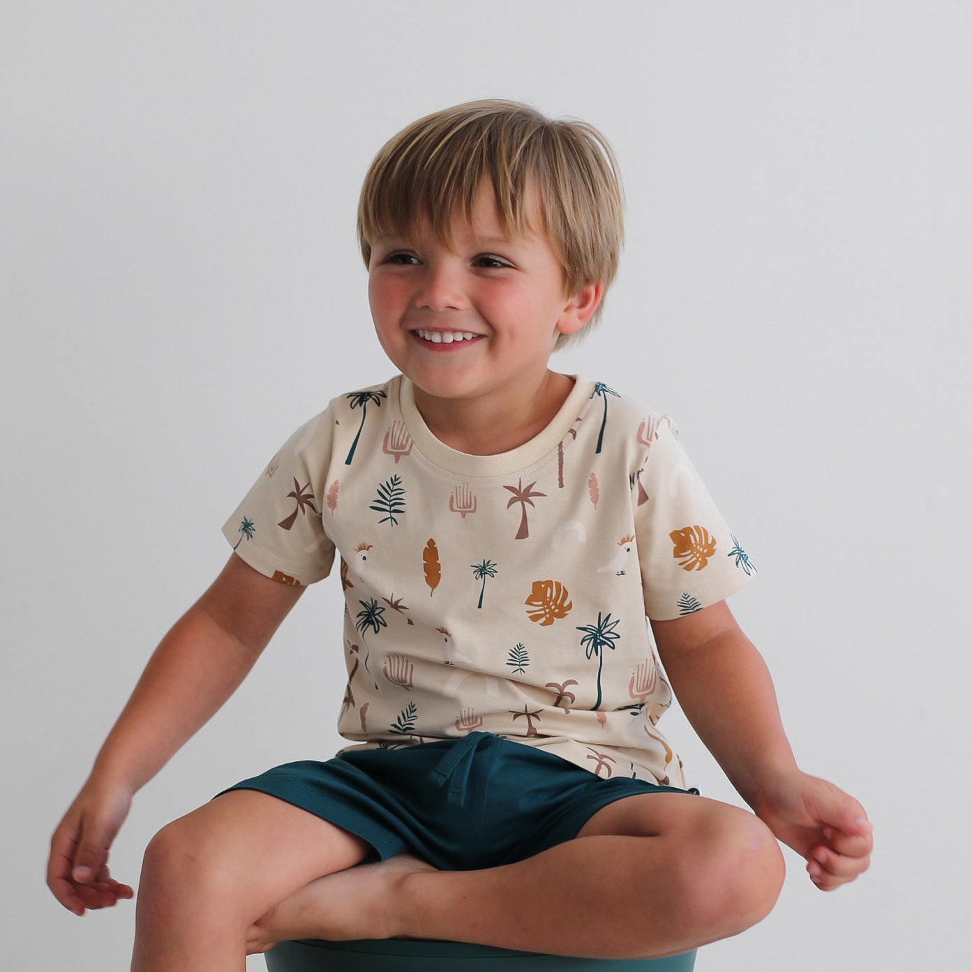 Boy sitting on stool while wearing Leo Shorts in Deep Teal and Jungle Stamp Classic T-Shirt, all designed by Burrow and Be