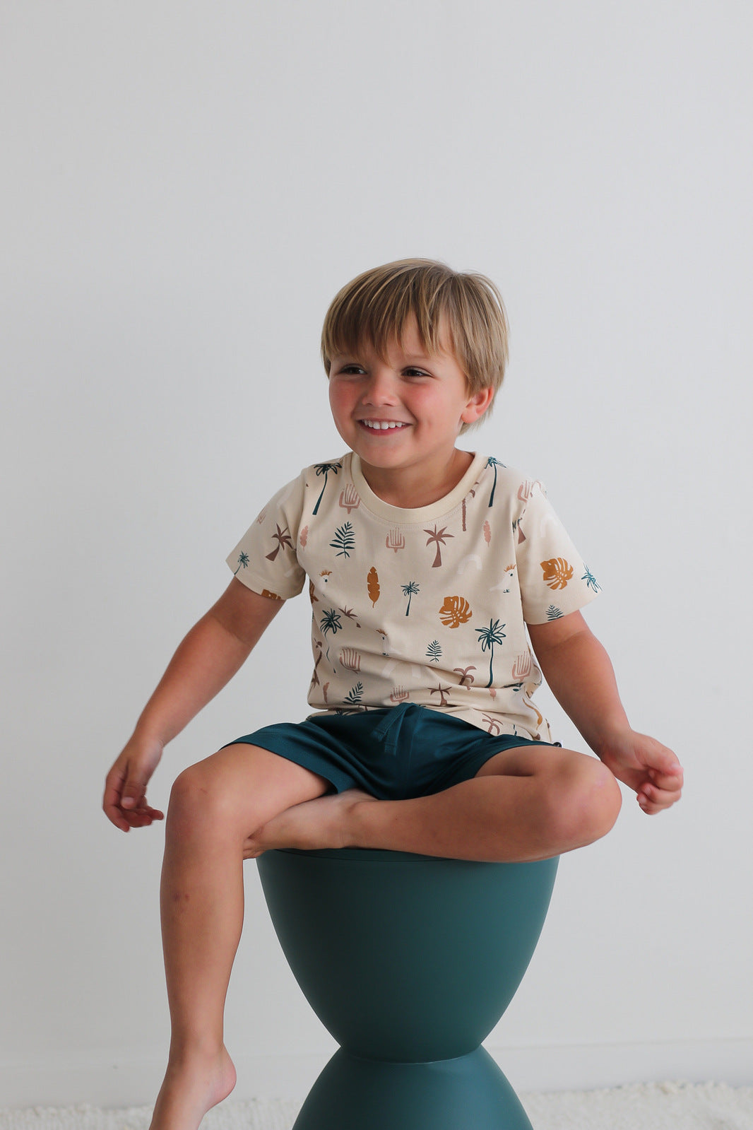 Boy sitting on a stool against a white wall while wearing Leo Shorts in Deep Teal and Jungle Stamp Classic T-Shirt, all designed by Burrow and Be