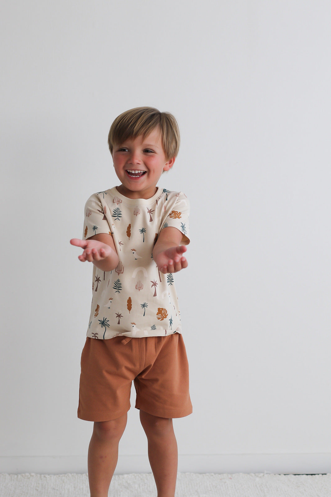 Boy posing against a white wall, standing on a white carpet while wearing Leo Shorts in Tan and Jungle Stamp Classic T-Shirt, all designed by Burrow and Be