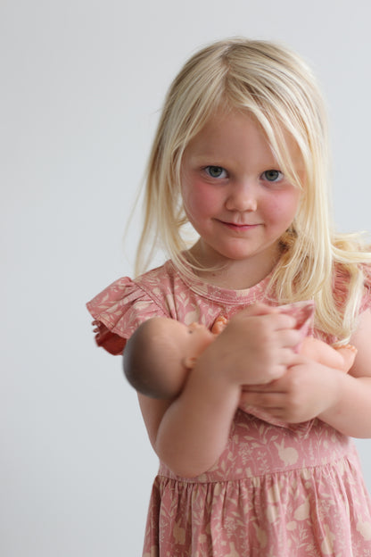 Girl wearing Mead-Doe Flutter Dress designed by Burrow and Be and holding a doll, posing in front of a white wall.