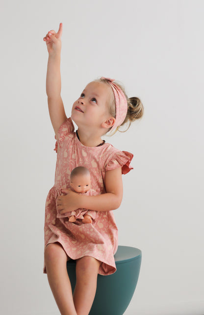 Girl sitting on a stool in front of a white wall, carrying a doll while wearing Mead-Doe Headband and Flutter Dress, all designed by Burrow and Be 