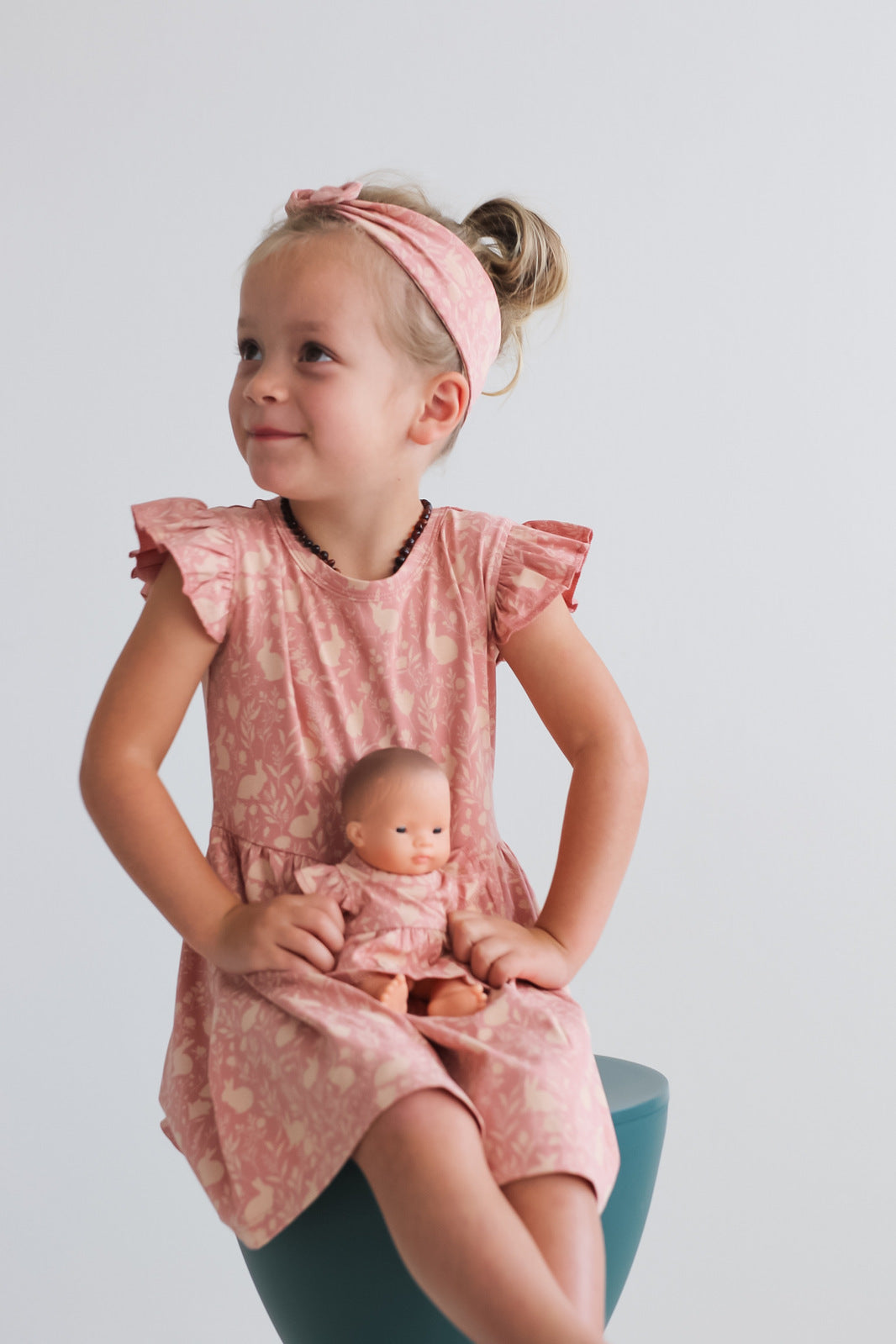 Girl posing and sitting on a stool in front of a white wall, carrying a doll while wearing Mead-Doe Headband and Flutter Dress, all designed by Burrow and Be 