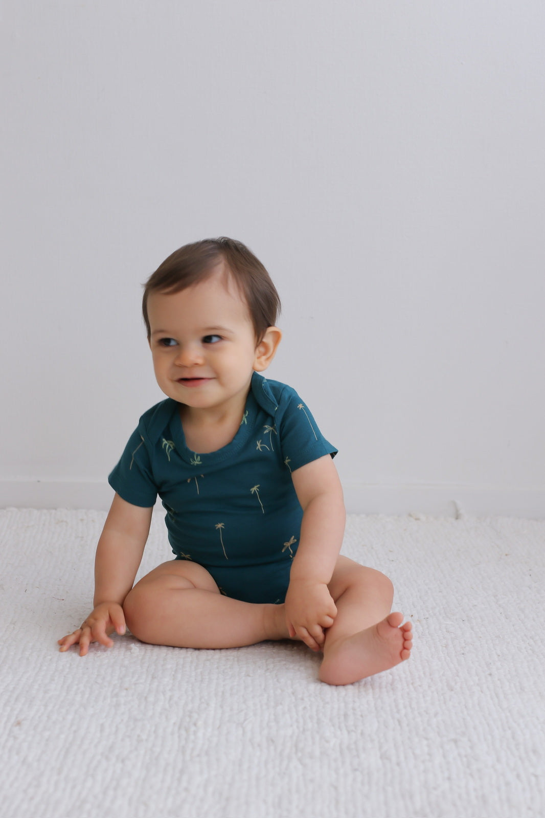 Baby sitting on white carpet while wearing Palm T-shirt Bodysuit, designed by Burrow and Be