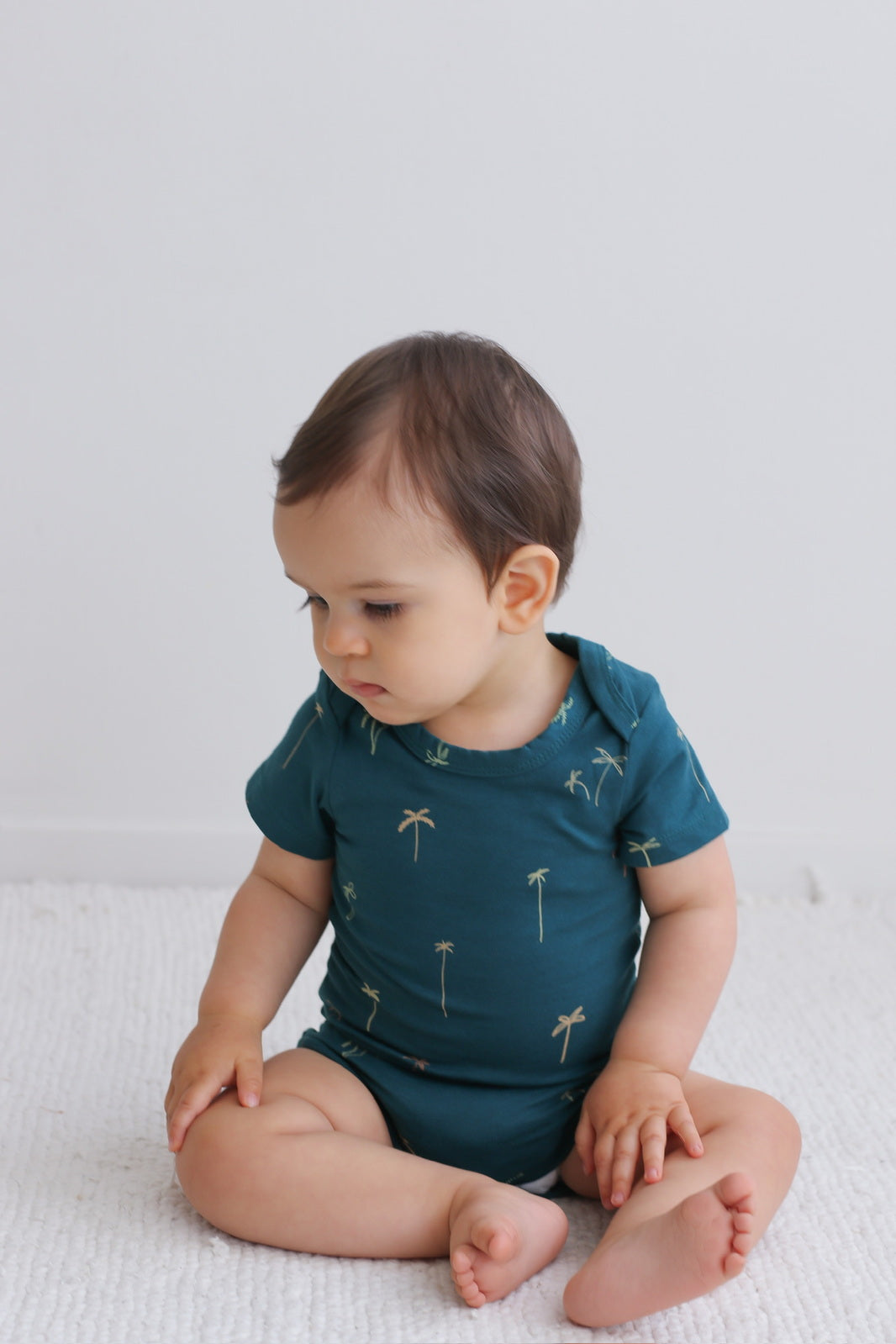 Baby sitting on white carpet in front of a white wall while wearing Palm T-shirt Bodysuit, designed by Burrow and Be