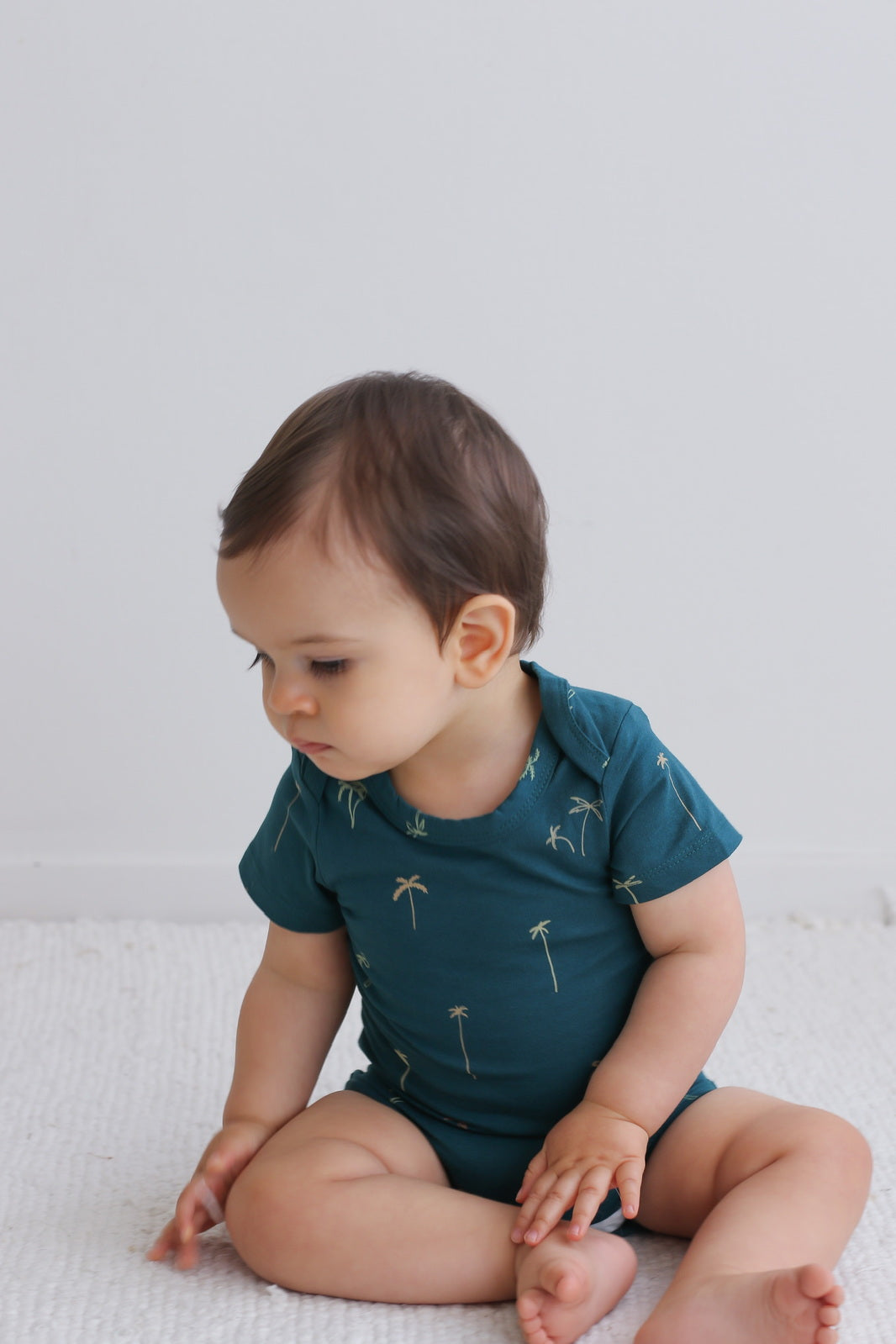 Baby sitting on white carpet while wearing Palm T-shirt Bodysuit, designed by Burrow and Be