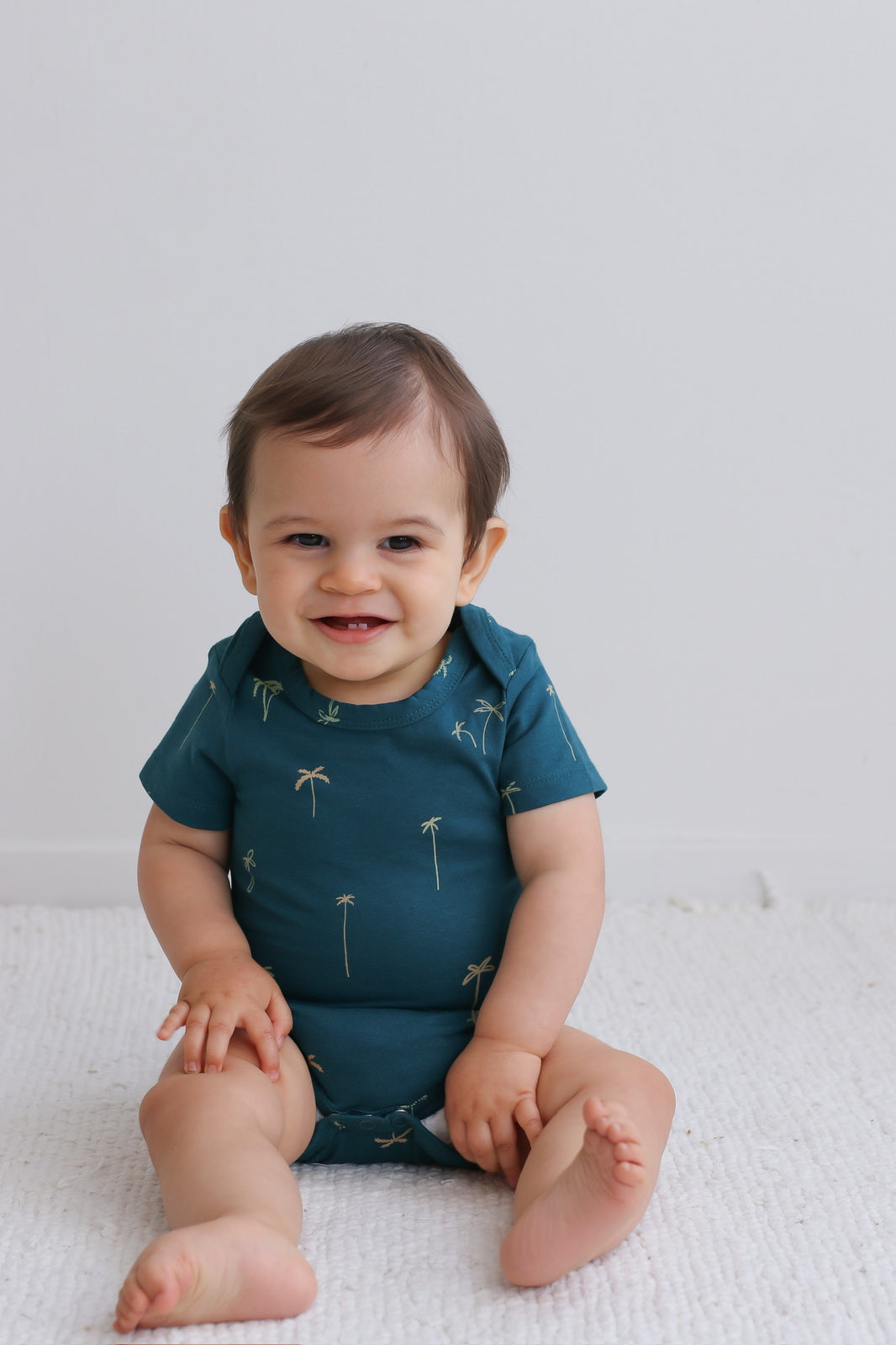 Baby sitting on white carpet while posing in Palm T-shirt Bodysuit, designed by Burrow and Be