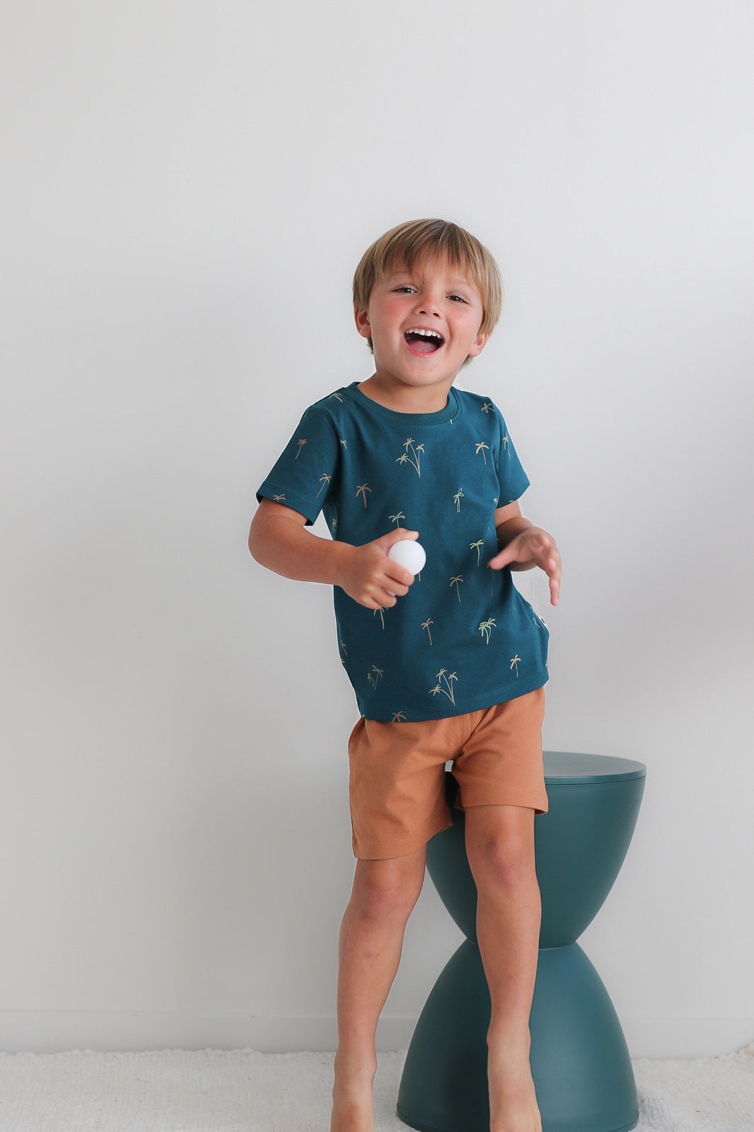 Boy holding a ball in front of a stool against a white background, wearing Palm Classic T-shirt and Leo Shorts in Tan, both designed by Burrow and Be