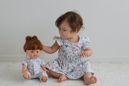 Girl sitting on a carpet in front of white background, sitting beside a baby doll dressed in Wildflower Doll Clothes while wearing Wildflower Flutter Baby Dress, both designed by Burrow and Be