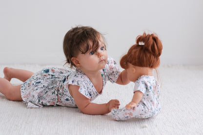 Girl laying on carpet in front of white background, playing with a baby doll dressed in Wildflower Doll Clothes while wearing Wildflower Flutter Baby Dress, both designed by Burrow and Be