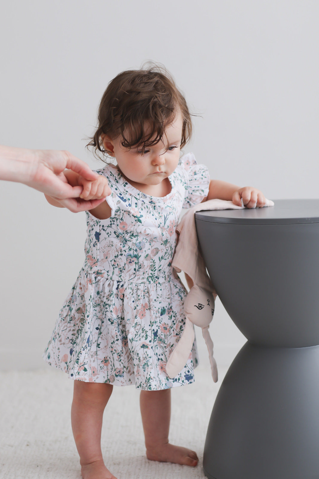 Girl posing in front of white background, beside a stool and wearing Wildflower Flutter Baby Dress, designed by Burrow and Be