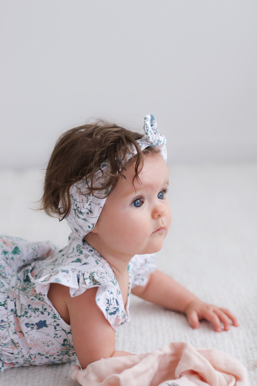 Girl sitting on a carpet in front of white background, while wearing Wildflower Headband and Flutter Baby Dress, both designed by Burrow and Be