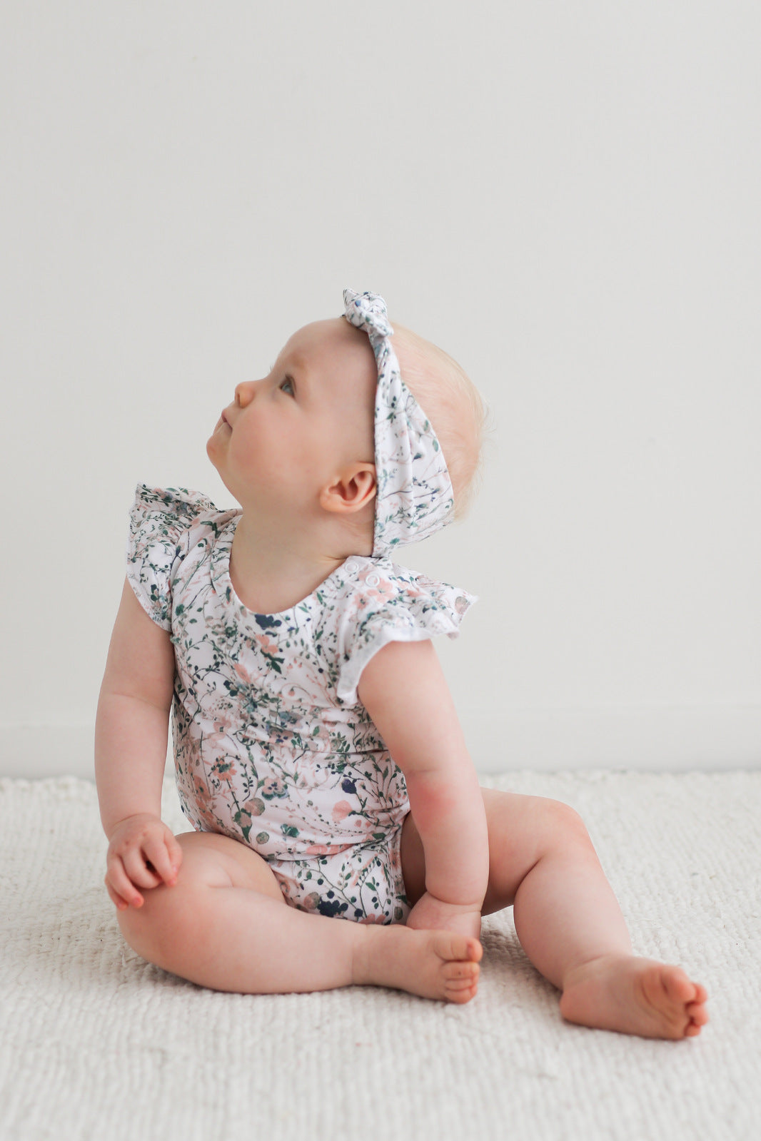 Baby sitting on carpet in front of a white wall, while wearing Wildflower Headband and Flutter Bodysuit, designed by Burrow and Be