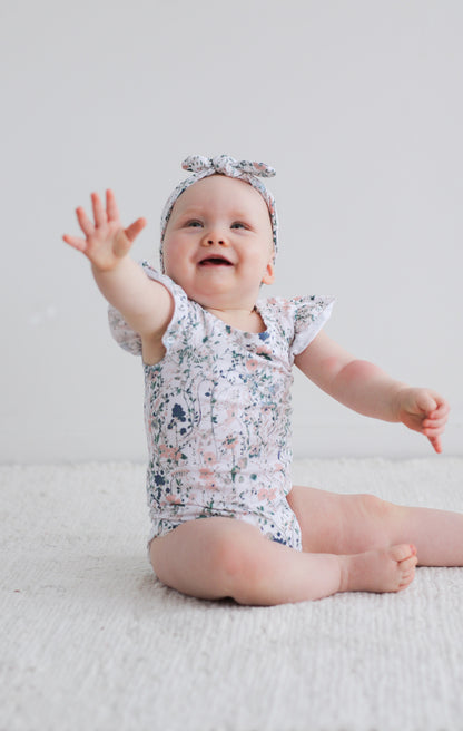 Baby sitting on carpet in front of a white wall, posing at the camera while wearing Wildflower Headband and Flutter Bodysuit, designed by Burrow and Be