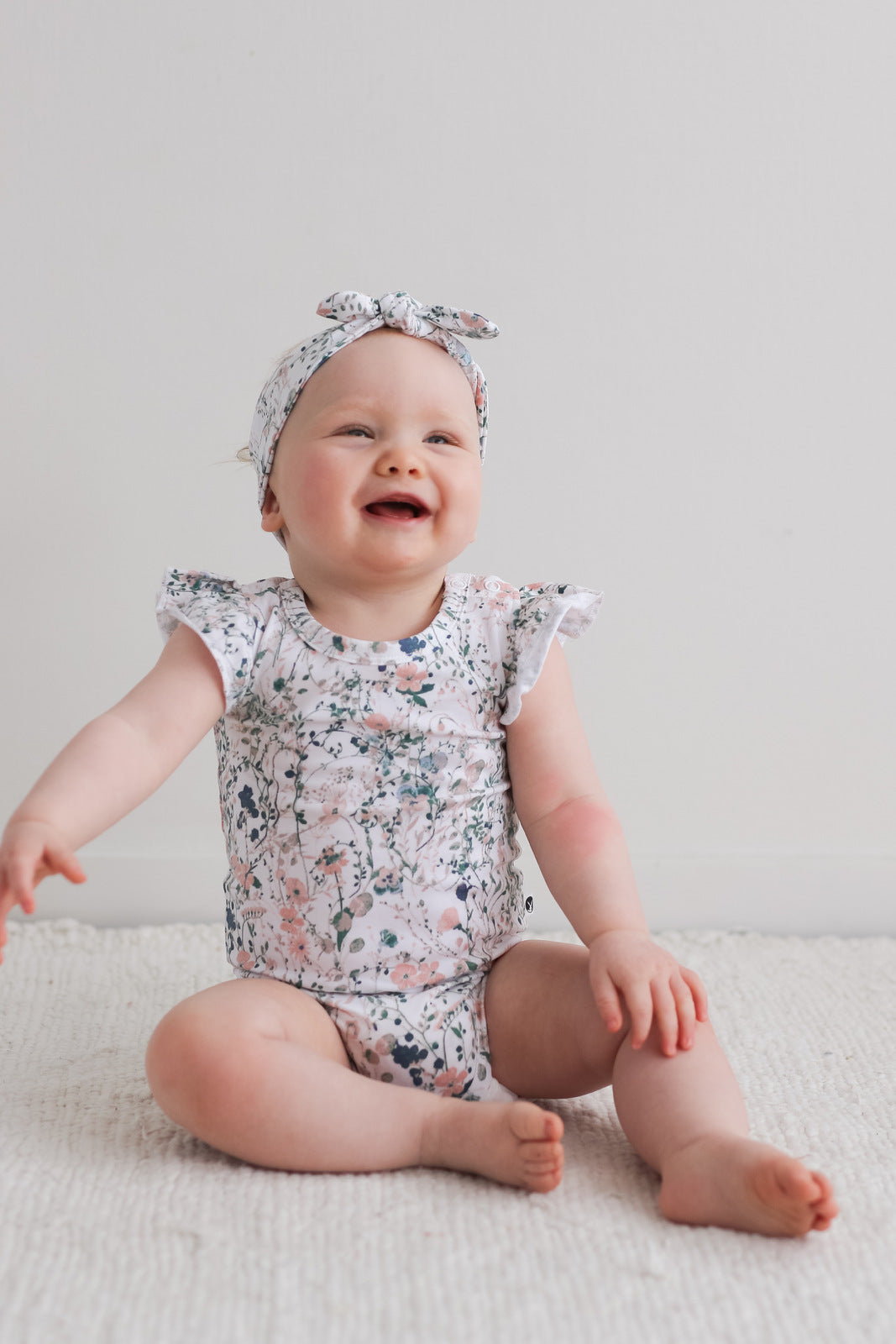 Baby sitting on carpet in front of a white wall, while wearing Wildflower Headband and Flutter Bodysuit, designed by Burrow and Be