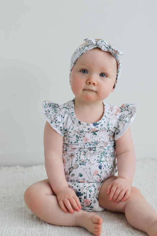 Baby sitting on carpet in front of a white wall, while wearing Wildflower Headband and Flutter Bodysuit, designed by Burrow and Be