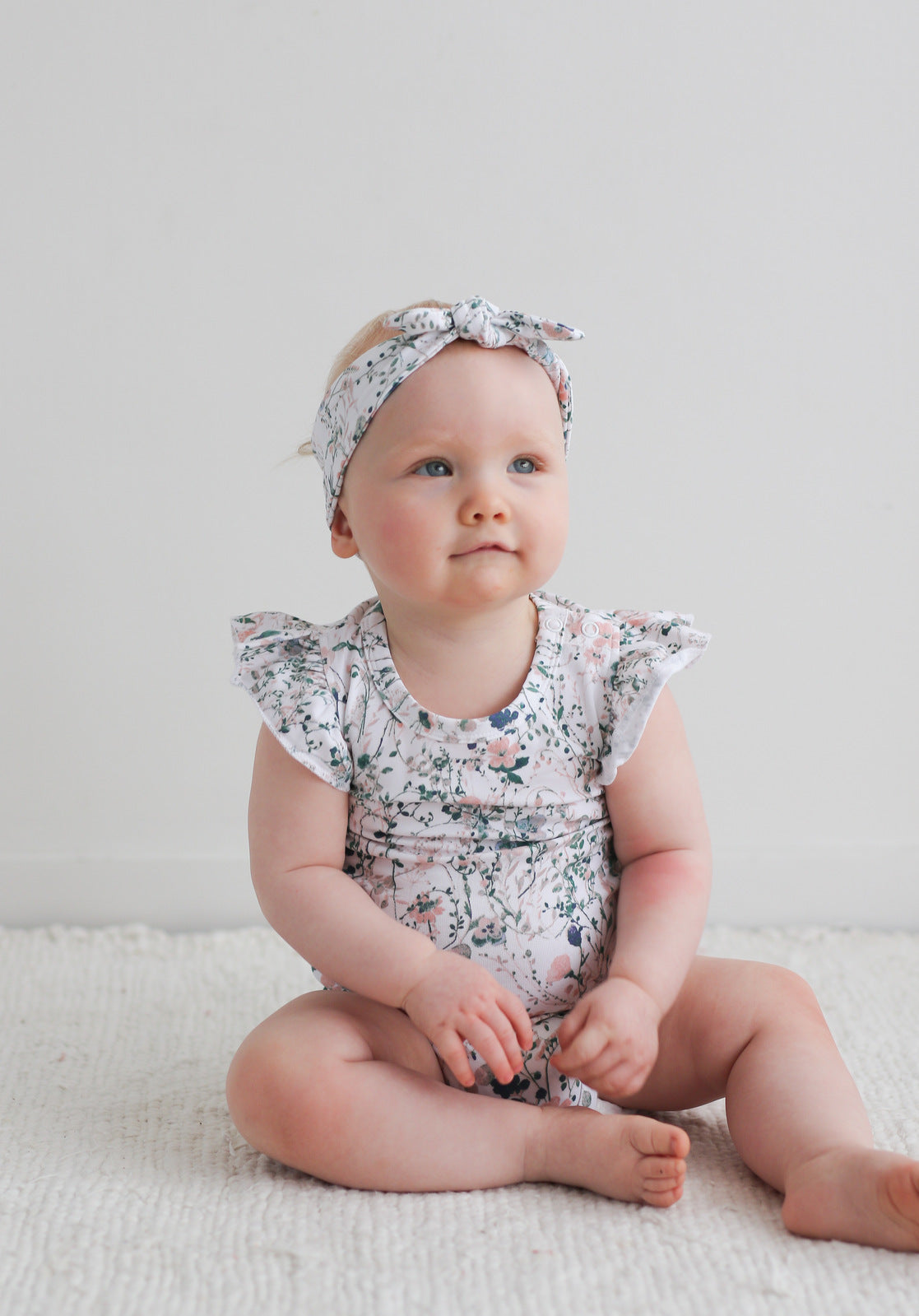 Baby sitting on carpet in front of a white wall, while wearing Wildflower Headband and Flutter Bodysuit, designed by Burrow and Be