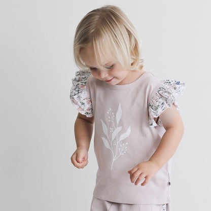 Girl standing in front of a white wall while wearing Wildflower/Lavender Flutter T-Shirt and Penny Shorts, both designed by Burrow and Be