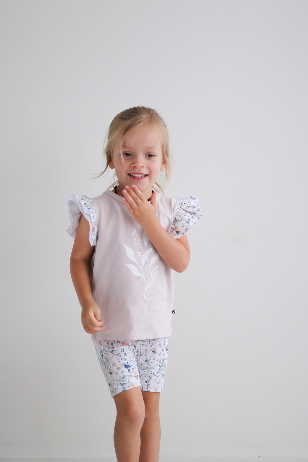 Girl standing in front of a white wall while wearing Wildflower/Lavender Flutter T-Shirt and Wildflower Bike Shorts, both designed by Burrow and Be