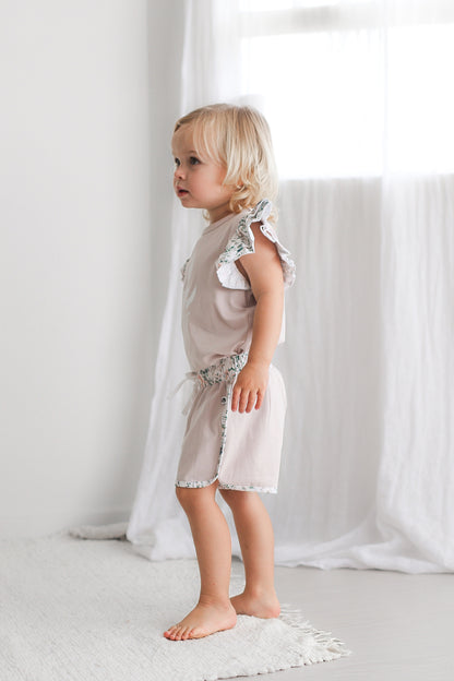 Girl standing in front of white curtains facing towards the right while wearing Wildflower/Lavender Flutter T-Shirt and Penny Shorts, both designed by Burrow and Be