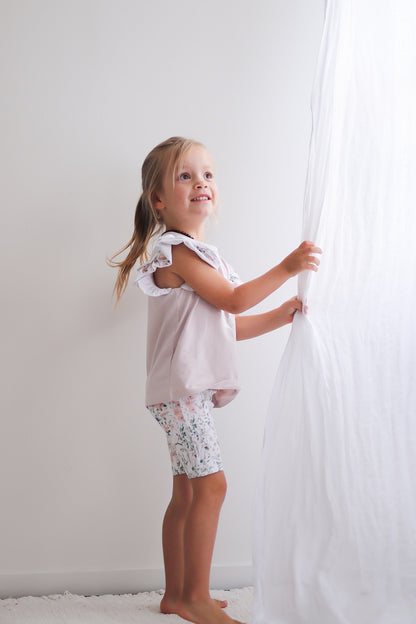 Girl standing in front of white curtains while wearing Wildflower/Lavender Flutter T-Shirt and Wildflower Bike Shorts, both designed by Burrow and Be