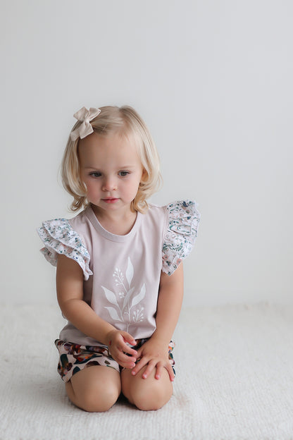 Girl sitting in front of a white wall while wearing Wildflower/Lavender Flutter T-Shirt and Garden Party Lyla Shorts, both designed by Burrow and Be