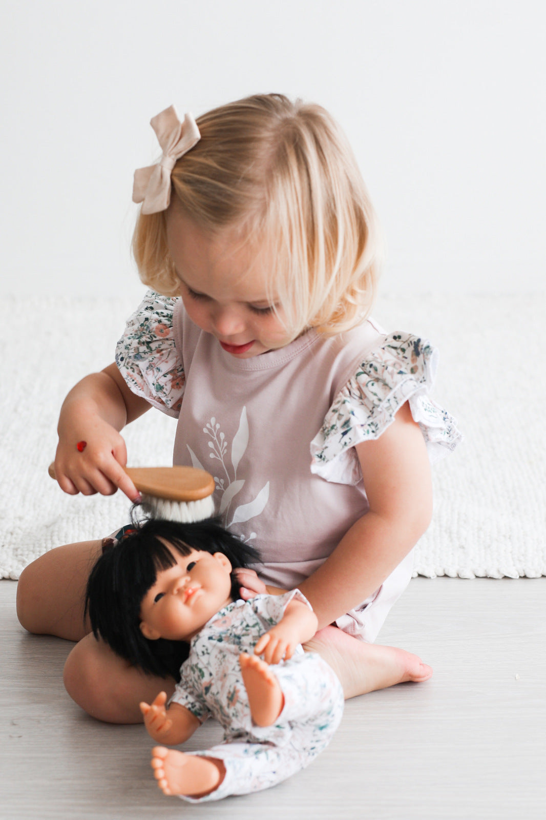 Girl sitting on the floor in front of carpet, brushing a doll that's wearing Wildflower Doll Clothes using Essential Baby Brush, while wearing Wildflower/Lavender Flutter T-Shirt and Penny Shorts, all designed by Burrow and Be