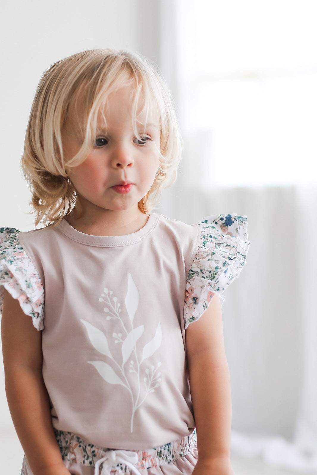 Girl standing in front of white curtains while wearing Wildflower/Lavender Flutter T-Shirt and Penny Shorts, both designed by Burrow and Be