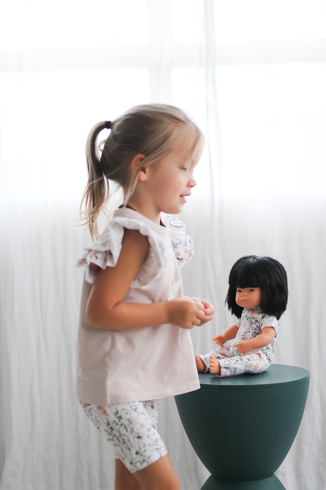 Girl standing in front of white curtains, playing with a doll dressed in Wildflower Doll Clothes seated on a stool while wearing Wildflower/Lavender Flutter T-Shirt and Wildflower Bike Shorts, both designed by Burrow and Bed