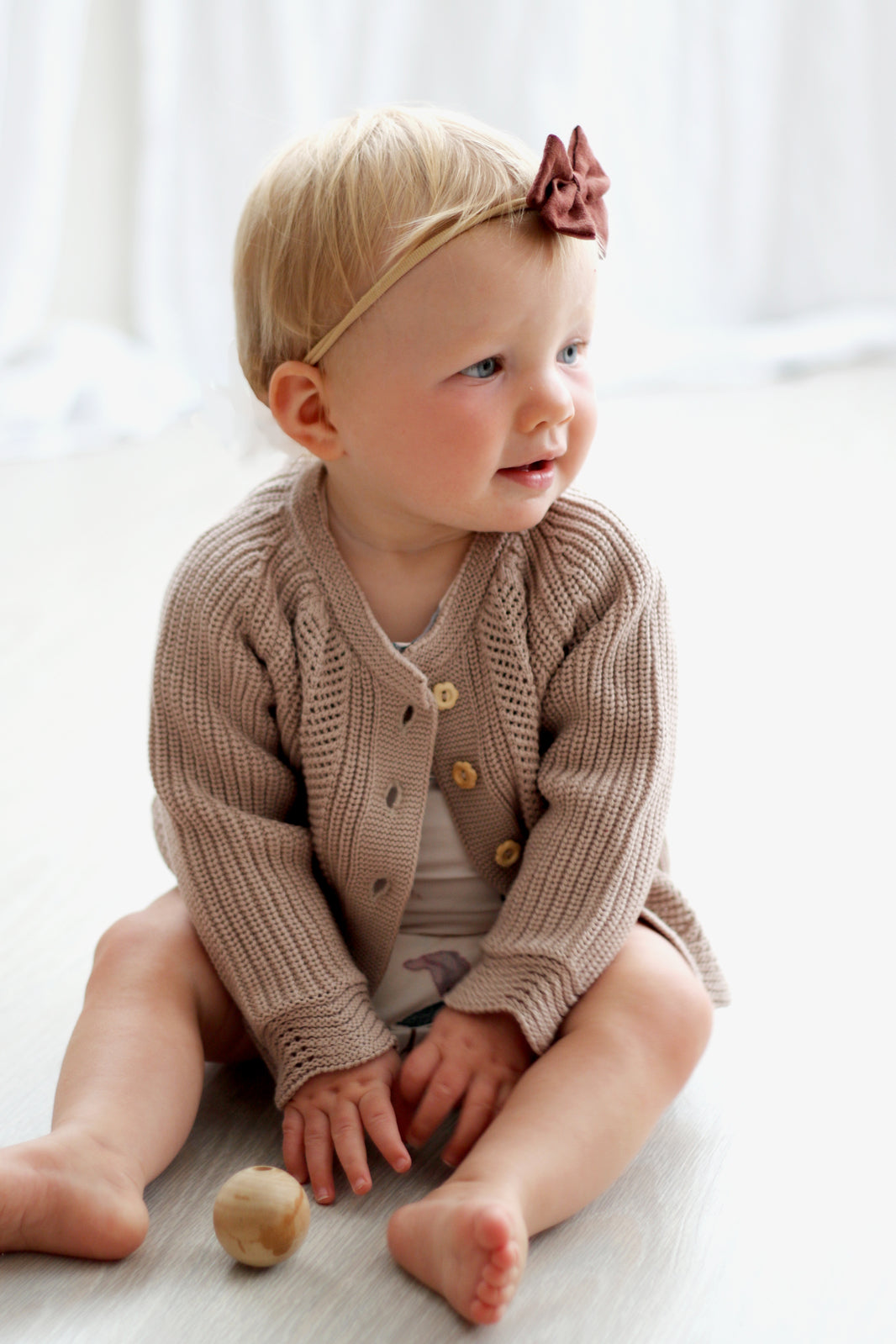 Baby girl sitting on a wooden floor and wearing Bow Tie hair band and wearing Florence Knit Cardigan, both designed by Burrow and Be