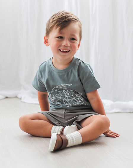 Boy sitting on wooden floor in front of white curtains, wearing Terry Knit Baby Shorts in Steel, Shoes by Wander and Lost on Storm T-Shirt, all designed by Burrow and Be